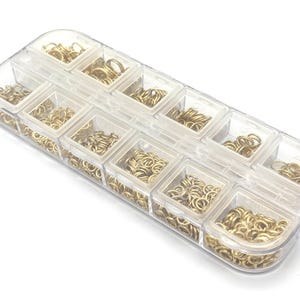 May include: A clear plastic organiser filled with small, gold-coloured jump rings. The rings are neatly arranged in individual compartments, suggesting a craft or jewellery-making supply. The container is rectangular with a curved top.