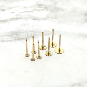 May include: A set of ten gold-coloured earring posts with flat backs. The posts are arranged in a semi-circle on a white surface.