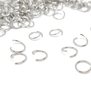 May include: A pile of silver jump rings, some are scattered on a white surface.