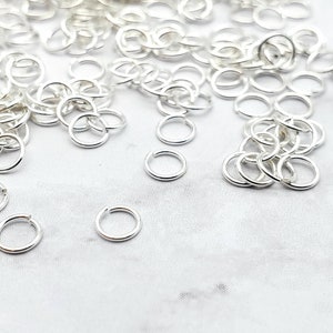 May include: A large pile of silver jump rings, a type of jewellery finding, on a white surface. The jump rings are small and round, and they are perfect for connecting different pieces of jewellery together.