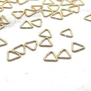 Op de afbeelding: Een groep gouden driehoekige sprongringen, verspreid over een wit oppervlak. Deze ringen zijn klein en delicaat, perfect voor het maken van sieraden.