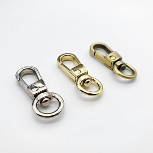 Könnte beinhalten: Drei Metall-Drehhaken in Silber, Gold und Antik-Messing. Jeder Haken hat einen federbelasteten Verschluss und einen kleinen Ring zur Befestigung an einem Riemen oder einer Kette. Die Haken befinden sich auf weißem Hintergrund.