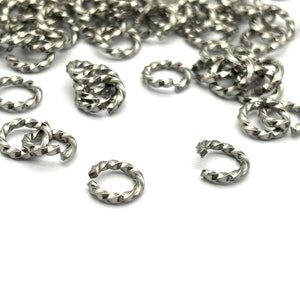 May include: A close-up of a pile of silver-coloured metal jump rings. The rings are twisted and have a diamond-shaped pattern.