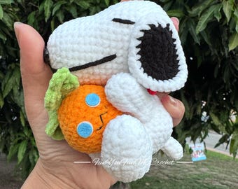 May include: A handmade crochet Snoopy plush toy in white with black ears and a red collar, holding an orange carrot with green leaves and blue eyes. The plush is made of soft yarn.