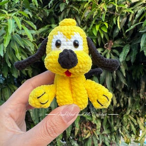 May include: A handmade yellow crochet dog with black ears and nose, and white accents around the eyes. The dog has a red tongue and is being held up in front of a green leafy background. The dog is a plush toy.