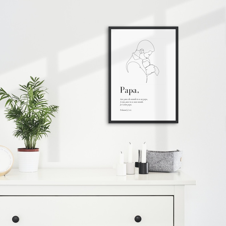 May include: Black and white line art print of a father holding a baby. The print is framed in a black frame and has the word "Papa" and a quote in French below the image.