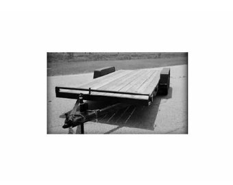 May include: A black trailer with a wooden deck and a single axle. The trailer has a metal railing around the edge of the deck.