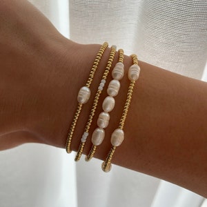 May include: A set of four gold beaded bracelets with white pearl accents. The bracelets are stacked on a person's wrist.