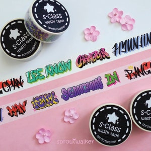 May include: Washi tape rolls with black and white labels featuring the text "s-class washi tape" and "sproutwalker". The tape itself displays colorful graffiti-style lettering with names. Pink paper flowers are scattered around.