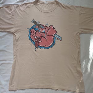 Könnte beinhalten: Ein beiges T-Shirt mit einem Cartoon-Schwein, das ein blaues und rotes Bandana trägt und ein Messer durch den Kopf hat. Das Schwein weint und hat einen blauen Stern auf der Nase.