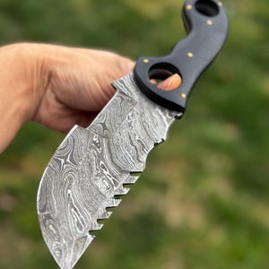 May include: A Damascus steel knife with a black wooden handle. The blade has a unique pattern and a serrated edge. The handle features two finger holes and gold-colored rivets.
