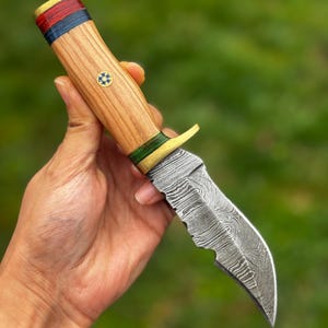 May include: A handcrafted knife with a wooden handle and a patterned steel blade. The handle features red, blue, and green accents, with a decorative emblem. The blade has a curved, serrated edge. The knife is held against a blurred green background.