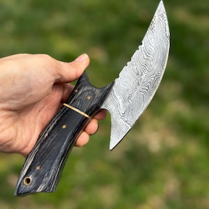 May include: A handcrafted knife with a curved, patterned blade and a dark wooden handle. The handle features brass accents and a gold band. The blade has a unique, swirling pattern. The knife is held against a blurred green background.