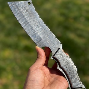 May include: A Damascus steel cleaver with a patterned blade and handle. The blade has a rectangular shape with a serrated edge and a hole near the top. The handle is intricately designed with a matching pattern.