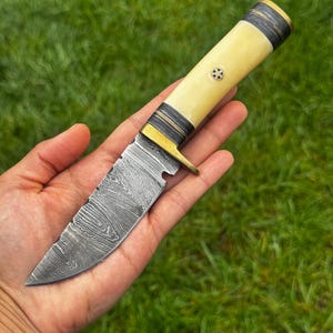 May include: A fixed-blade knife featuring a patterned Damascus steel blade with a distinct wavy texture. The handle is crafted from a smooth, cream-colored material with dark, layered spacers, a brass bolster, and a decorative mosaic pin inlay.