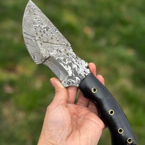 May include: A Damascus steel knife with a patterned blade and a black handle. The handle is secured with three brass rivets. The knife is held against a blurred green background.