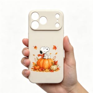 May include: A cream-colored smartphone case featuring a graphic of the cartoon character Snoopy wearing a red and white plaid scarf, sitting on a large orange pumpkin surrounded by smaller pumpkins and scattered autumn leaves in shades of orange and red.