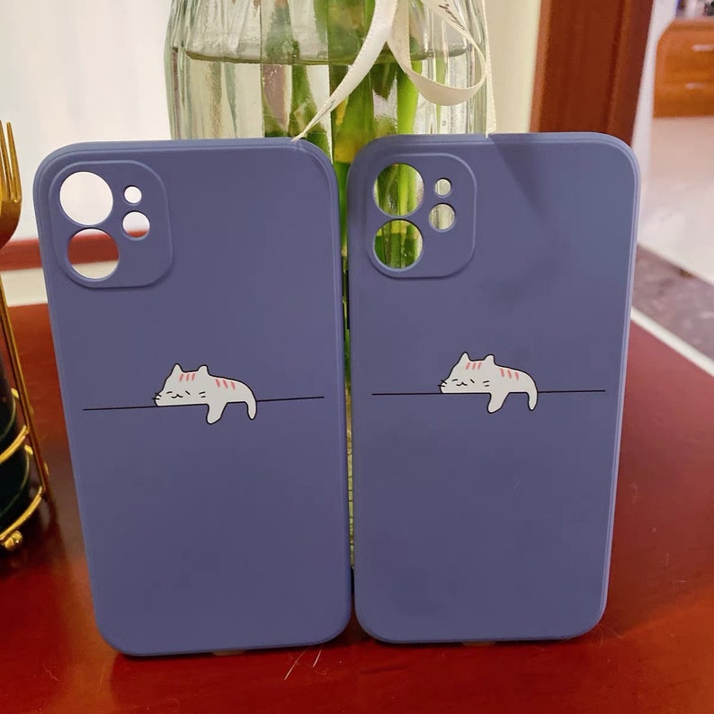 May include: Two purple phone cases with a white cat design. The cat is lying down with its head resting on a line. The cases have a camera cutout.