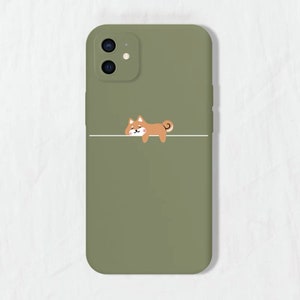 May include: A green phone case featuring a cartoon Shiba Inu dog illustration. The dog is brown and white and is lying down on a white line.