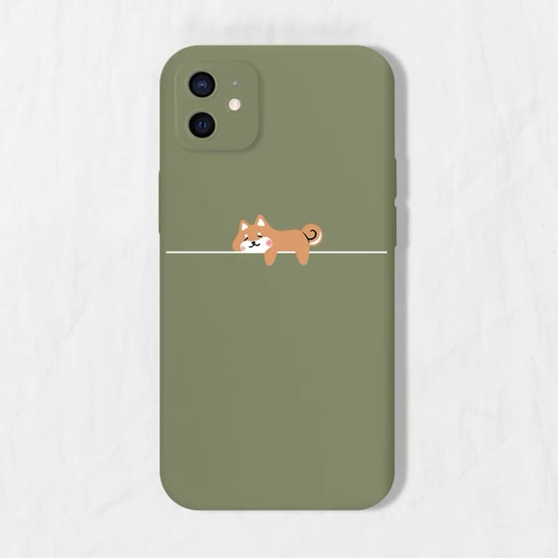 May include: A green phone case featuring a cartoon Shiba Inu dog illustration. The dog is brown and white and is lying down on a white line.