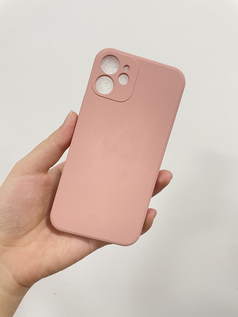 Peut inclure: Une coque de t&eacute;l&eacute;phone rose poudr&eacute; avec des ouvertures pour les objectifs de l'appareil photo. L'&eacute;tui est de couleur unie et semble &ecirc;tre fait d'un mat&eacute;riau lisse. L'&eacute;tui du t&eacute;l&eacute;phone est tenu dans une main sur fond blanc.