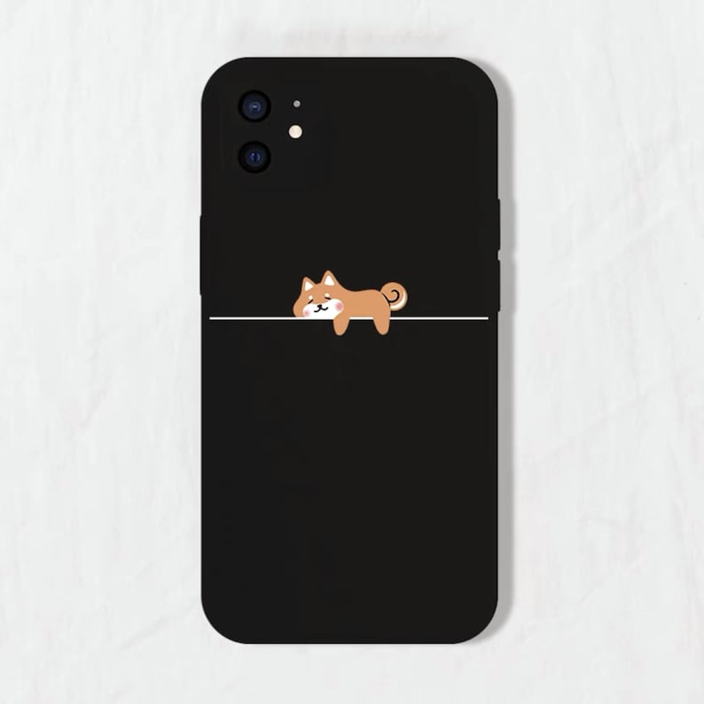 May include: Black phone case featuring a cute cartoon Shiba Inu dog illustration. The dog is brown and white and is lying down with its head resting on a white line.