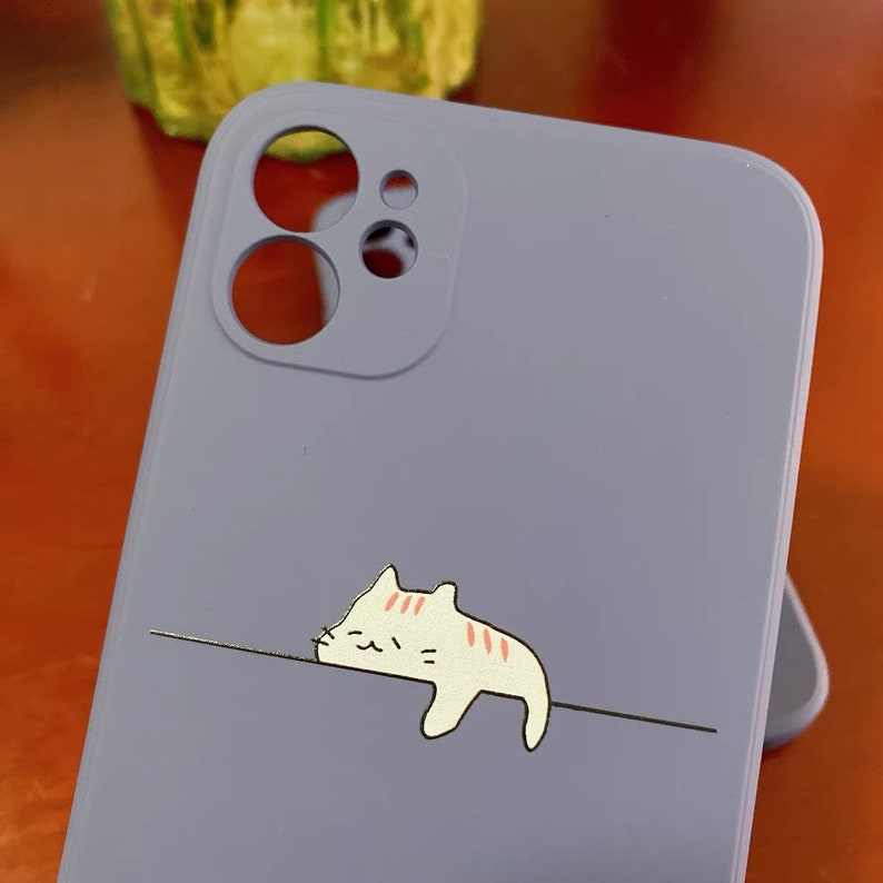 May include: A light purple phone case with a cartoon cat design. The cat is white with pink stripes and is lying down on a line.