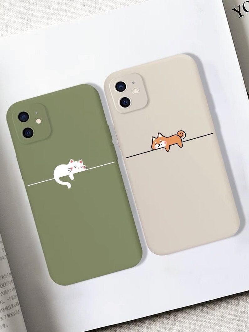 May include: Two phone cases featuring cute animal illustrations. The case on the left is green with a white cat sleeping on a line. The case on the right is beige with an orange dog sleeping on a line.