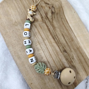 May include: A beige baby pacifier with a beaded chain. The chain features giraffe and leaf-shaped beads, along with lettered beads spelling "LOHAN". The pacifier is attached to a metal clip.