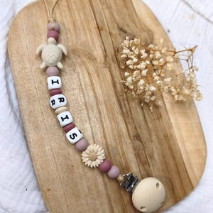 Pode incluir: Um clipe de chupeta para bebé com o nome "IRIS" escrito em contas de letras. O clipe apresenta um encanto de tartaruga bege, contas rosa e malva, e um encanto de flor. A extremidade da chupeta é de cor bege claro.