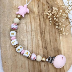 Könnte beinhalten: Ein Schnullerhalter mit einer rosa Silikonschildkröte, goldenen, cremefarbenen und blumig gemusterten Perlen sowie weißen Würfelperlen, die den Namen ELEA in goldenen Buchstaben bilden. Das Accessoire hat eine weiße Kordelschlaufe und einen rosa Clip.