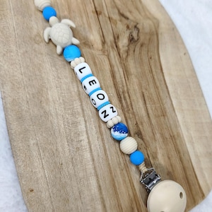 Könnte beinhalten: Ein Baby-Schnullerclip mit dem Namen "LEON" in weißen Buchstabenperlen. Der Clip hat blaue und Holzperlen, einen weißen Schildkrötenanhänger und einen beigen Schnullerhalter. Der Clip ist an einem Holzbrett befestigt.