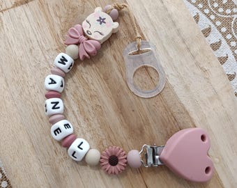 May include: A pacifier clip featuring a string of beads in shades of dusty rose, beige, and white. The beads spell the name MANELL and include a bear-shaped bead with a bow, a sunflower-shaped bead, and a heart-shaped clip in dusty rose.