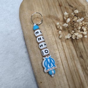 May include: A keychain with the name "ENZO" spelled out in white letter beads. The keychain also features a blue and white emblem with the text "DROIT AU BUT". The keychain is attached to a silver keyring.