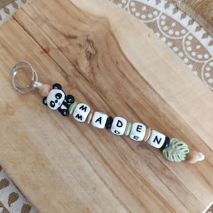 Peut inclure: Un porte-clés avec un charme panda noir et blanc avec le nom "MADEN" écrit en lettres noires sur des perles blanches. Les perles sont entrecoupées de perles brunes et vertes.
