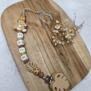 Könnte beinhalten: Ein Baby-Schnullerclip mit dem Namen "ZACK" in Holzperlen geschrieben. Der Clip zeigt ein Löwenjunges, Blätter und Perlen mit Giraffenmuster. Der Clip ist an einem durchsichtigen Kunststoffring und einem blattförmigen Holzstück befestigt.