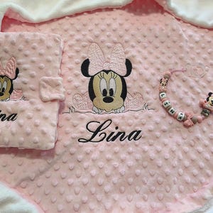 以下が含まれることがあります： ピンク色のベビーセット。ミニーマウスの刺繍と「Lina」の名前が入ったブランケット、おむつ替えパッド、おしゃぶりクリップのセット。ブランケットとパッドは、テクスチャード加工されたドットパターンです。