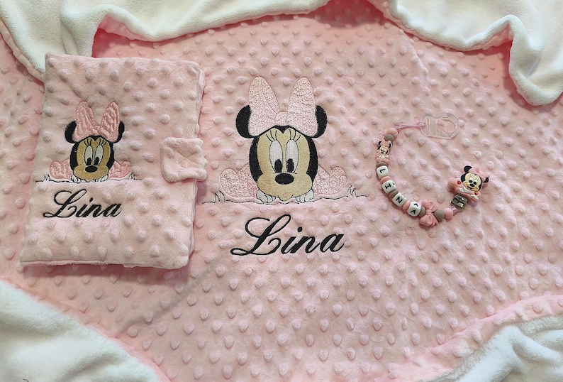 以下が含まれることがあります： ピンク色のベビーセット。ミニーマウスの刺繍と「Lina」の名前が入ったブランケット、おむつ替えパッド、おしゃぶりクリップのセット。ブランケットとパッドは、テクスチャード加工されたドットパターンです。