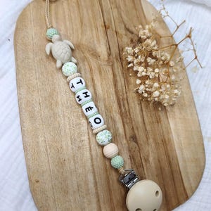 Peut inclure: Une attache-sucette composée de perles dans des tons vert sauge, crème et blanc. Les perles incluent une forme de tortue en silicone et des lettres formant le prénom THEO. Une pince métallique est fixée à une sucette ronde beige.