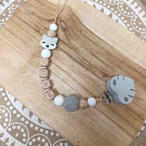 May include: A wooden and silicone baby teether with a fox charm and the name "NOAM" spelled out in wooden beads. The teether has a gray crocheted ball and a gray silicone leaf-shaped clip.