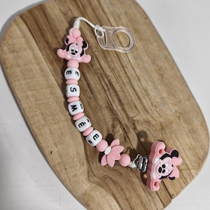 Può includere: Una catenella portaciuccio con un personaggio di Minnie in silicone rosa a ciascuna estremità, collegata da perline sferiche bianche che compongono il nome ESMÉE in lettere nere, alternate a piccole perline rosa e un ciondolo a forma di fiore.