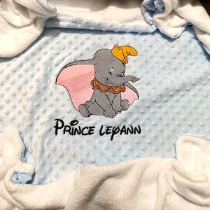 Puede incluir: Una manta de bebé azul claro con un borde blanco. La manta tiene una imagen bordada de Dumbo el elefante con el texto "Prince Lepann".
