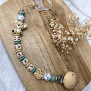 Può includere: Un porta ciuccio personalizzato per bambini con il nome "HANNA" scritto con perline di legno e verde acqua. La clip presenta un koala, una foglia e perline decorate. L'estremità del ciuccio è color beige chiaro.