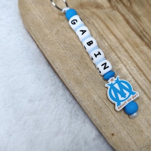 May include: A keychain with the name "GABIN" spelled out in white letter beads and blue accents. The keychain features a silver ring and a blue and white emblem with the text "DROIT AU BUT".