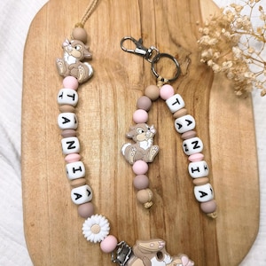 Op de afbeelding: Een speenkoord en een bijpassende sleutelhanger met siliconen kralen in tinten beige, bruin en roze. Beide items bevatten een konijnfiguurtje, een witte madeliefjeskraal en witte letterkralen die de naam TANIA vormen.
