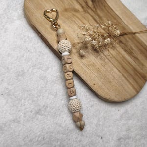 May include: A wooden keychain with the name "LOUIS" spelled out in wooden letter beads. The keychain features a gold heart-shaped clasp, crochet beads, and geometric wooden beads. The background includes a wooden cutting board and dried flowers.