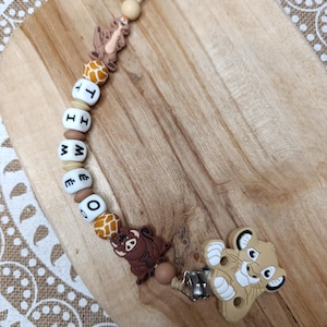 May include: A brown and white baby teether with a lion, a zebra, and a giraffe. The teether has a wooden clip and the letters "TIMEO" spelled out on it.