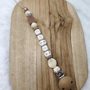 May include: A baby's pacifier clip with the name "JULIA" spelled out in white letter beads with gold letters. The clip features a brown pacifier, wooden beads, and a leaf-shaped bead. The clip is attached to a wooden board.