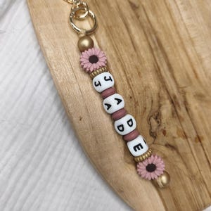 Peut inclure: Un porte-clés doré avec un fermoir en forme de cœur. Le porte-clés comporte des perles de lettres blanches épelant "JADE", entrecoupées de fleurs roses et de perles dorées. Le porte-clés est sur une surface en bois.