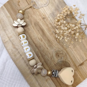 Pode incluir: Um prendedor de chupeta com uma série de contas em tons de dourado, bege e creme. As contas incluem dois detalhes em forma de flor e contas de letras brancas que formam o nome MILA em dourado. Um clipe bege em forma de coração está em uma ponta.